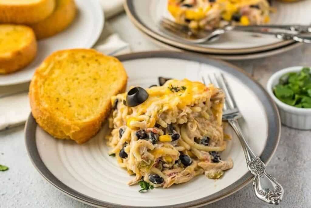 A plate with a serving of cheesy pasta bake&mdash;one of our favorite casserole recipes&mdash;containing black beans, corn, and olives, accompanied by a slice of garlic bread.