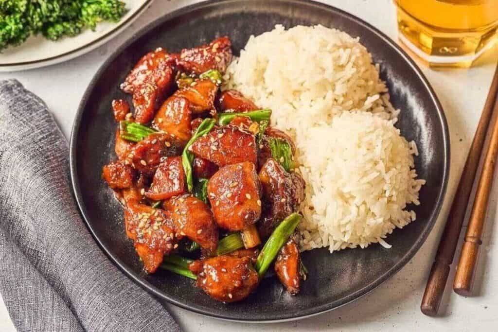 A black plate with glazed chicken pieces, green onions, and sesame seeds sits next to two servings of white rice&mdash;perfect for easy Weeknight Dinners. Chopsticks and a glass of drink are nearby.