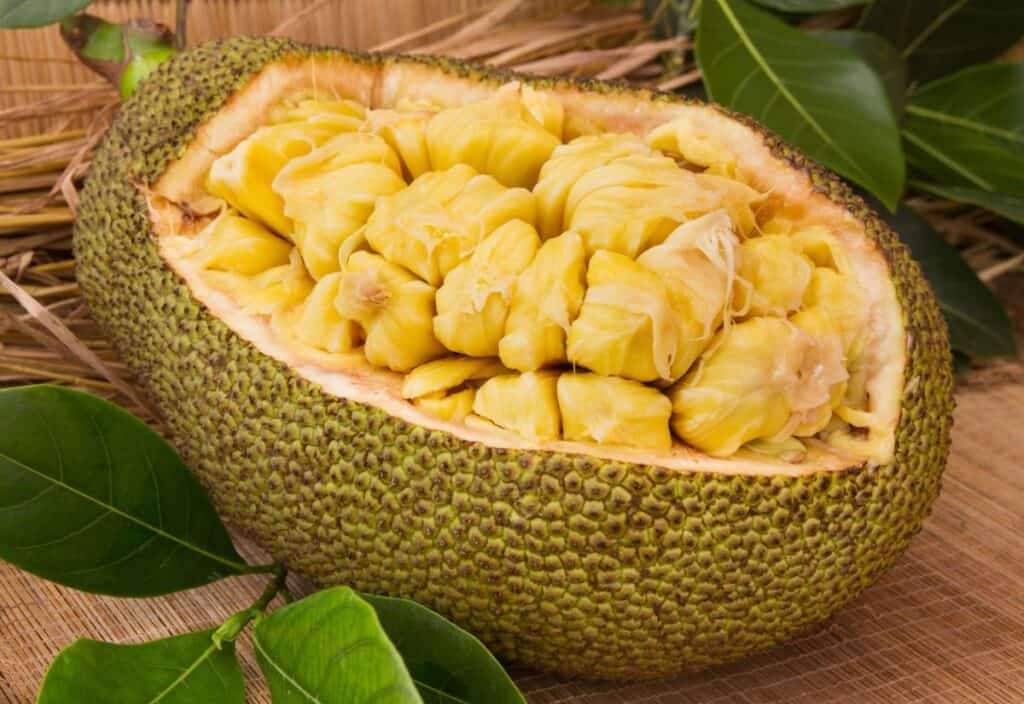 A large, green jackfruit cut in half, showing its yellow, segmented edible flesh and seeds, with green leaves and straw in the background.