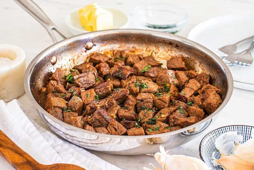 A stainless steel skillet filled with cooked, cubed steak garnished with herbs sits on a white surface&mdash;perfect for serving as delicious Game Day Snacks alongside butter, plates, and utensils.