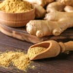 A wooden scoop and bowl contain ground ginger powder, with whole ginger roots placed on a wooden surface in the background.