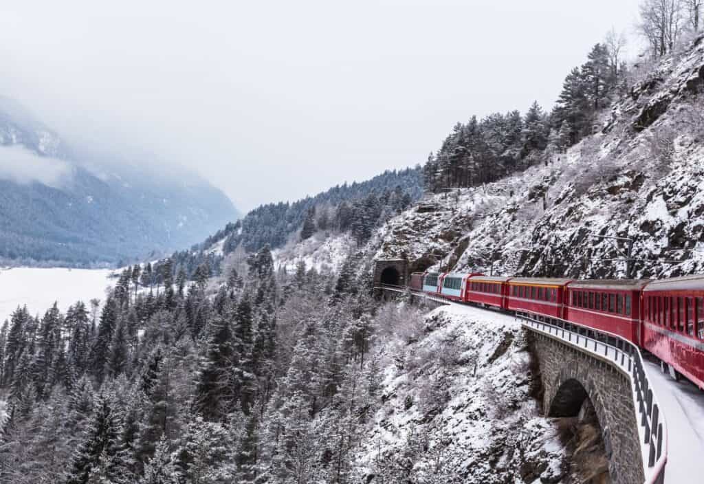 A red train travels along a curved railway track through a snowy mountain landscape lined with pine trees and tunnels.
