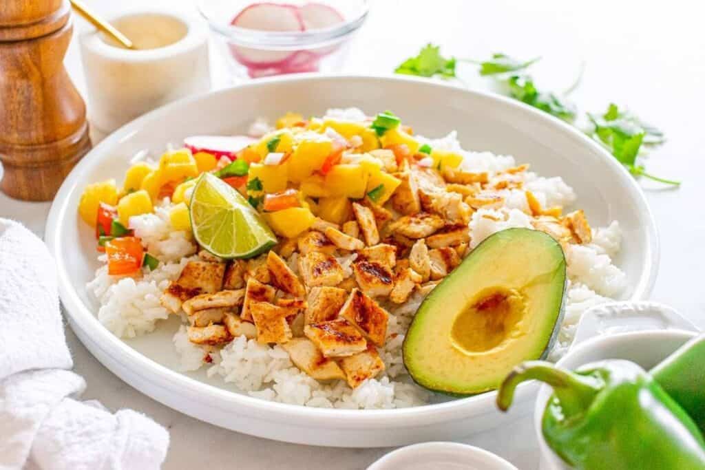 A perfect choice for weeknight dinners, this plate features white rice topped with diced grilled chicken, mango salsa, a lime wedge, and half an avocado, garnished with cilantro and a pepper on the side.