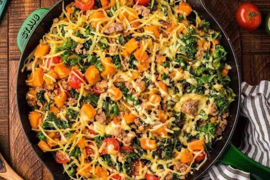 A cast iron skillet filled with a hearty weeknight dinner of ground meat, kale, diced sweet potatoes, cherry tomatoes, and melted shredded cheese.