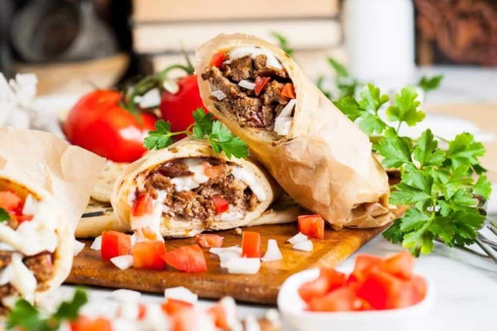 A close-up of a sliced wrap filled with grilled meat, diced tomatoes, onions, and sauce, placed on a wooden board with fresh parsley and chopped vegetables&mdash;perfect for delicious Game Day Snacks.