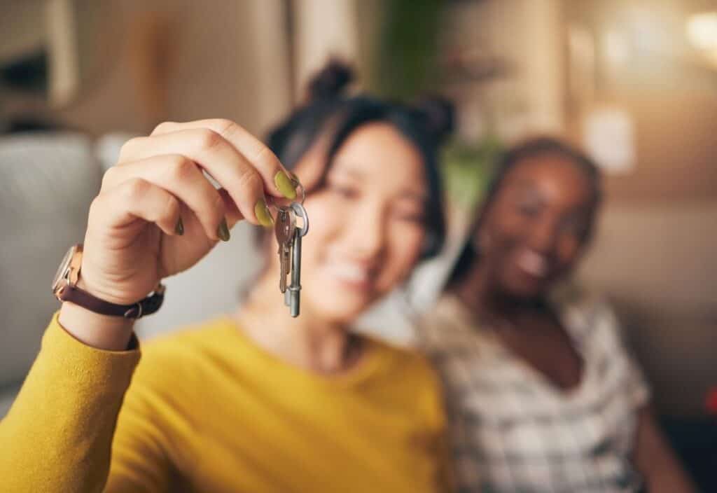A person in a yellow top holds a key toward the camera, with another person smiling beside them in the background&mdash;perfectly capturing the excitement of house hacking.