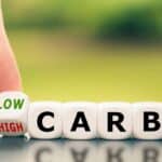 A hand flips a die showing "LOW" and "HIGH" beside blocks spelling "CARB," symbolizing carb choices&mdash;perfect for celebrating National Keto Day.