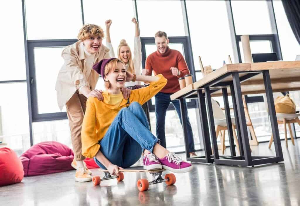 Four young adults celebrate National Fun at Work Day in a bright room; one sits on a skateboard while another pushes, and two others cheer nearby by a table and beanbags.