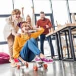 Four young adults celebrate National Fun at Work Day in a bright room; one sits on a skateboard while another pushes, and two others cheer nearby by a table and beanbags.