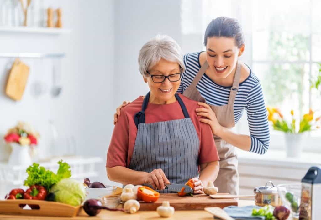 An older woman, a true grandfluencer, chops vegetables on a cutting board in the kitchen while a younger woman in an apron stands behind her, smiling warmly.