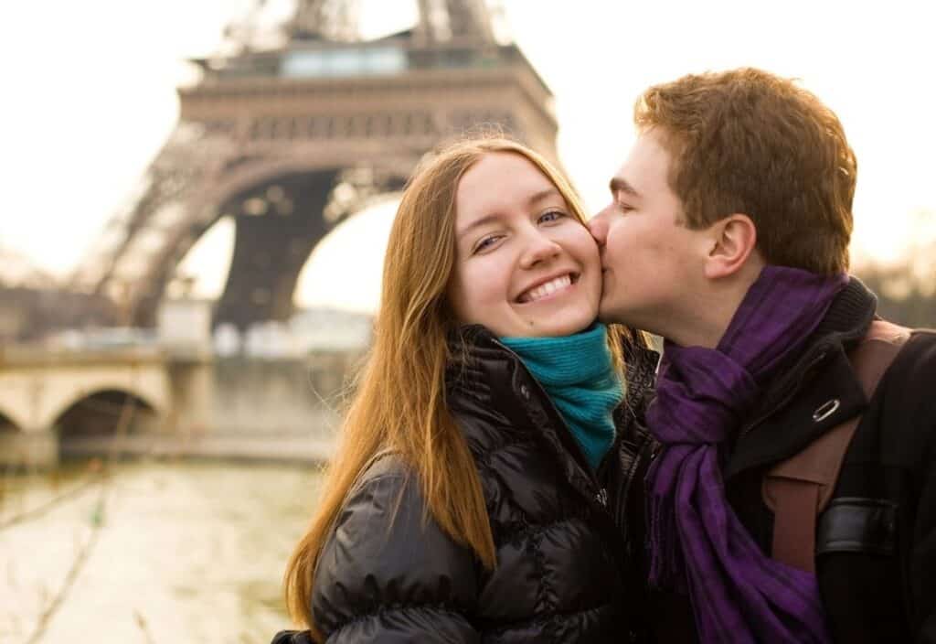 A couple enjoys romantic travel in Paris, posing in front of the Eiffel Tower; the man kisses the smiling woman on the cheek as they both wear winter jackets and scarves.