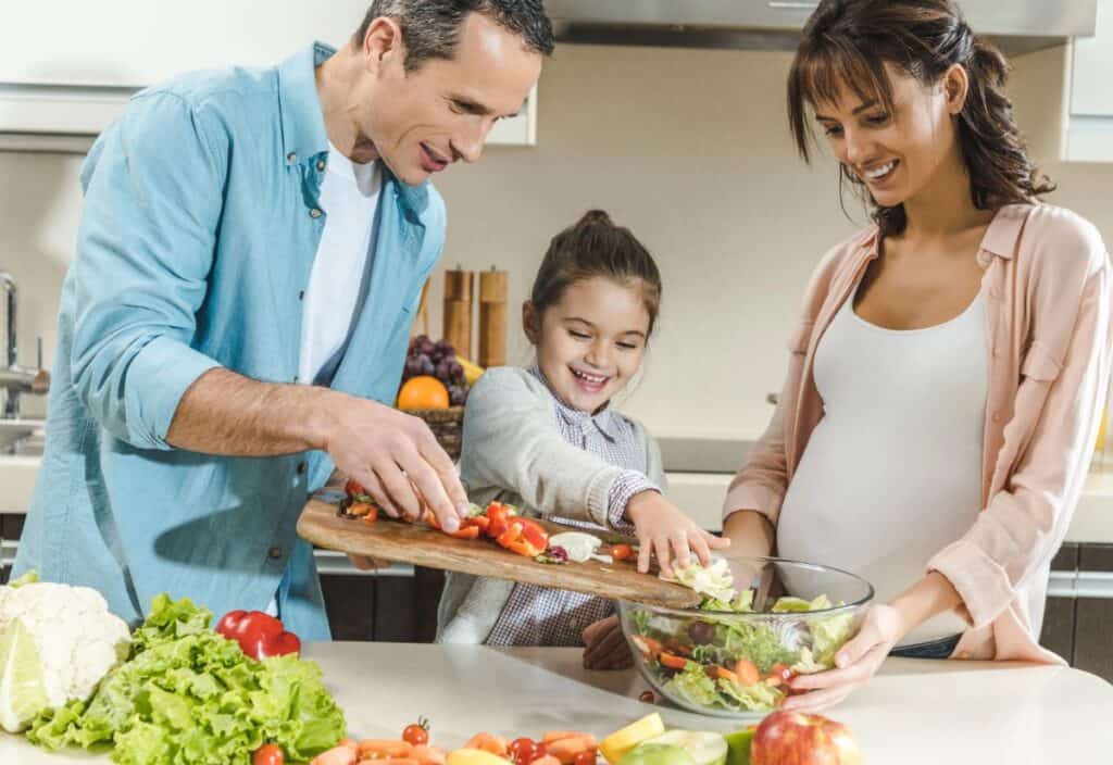 A man, woman, and child stand in a kitchen preparing a salad together, smiling as they add chopped vegetables to a glass bowl on the counter.