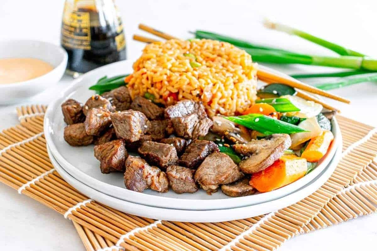 A plate with grilled steak bites, fried rice, and sautéed vegetables sits on a bamboo mat with chopsticks—an ideal choice for low effort dinners, complete with sauce and soy sauce in the background.