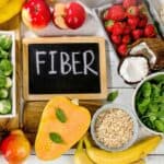 A variety of high-fiber foods including brussels sprouts, apples, strawberries, bread, spinach, oats, squash, bananas, pears, and coconut surround a chalkboard labeled "FIBER".