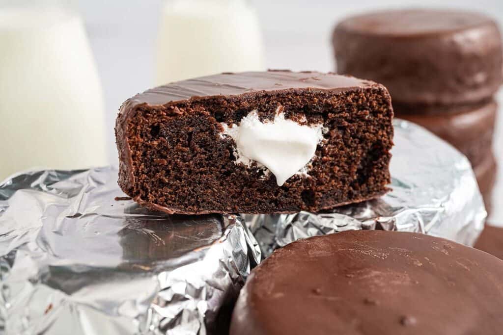 A chocolate snack cake with a creamy filling is cut in half, displayed on top of foil-wrapped cakes, with bottles of milk in the background&mdash;perfect for anyone seeking good desserts to satisfy a sweet craving.
