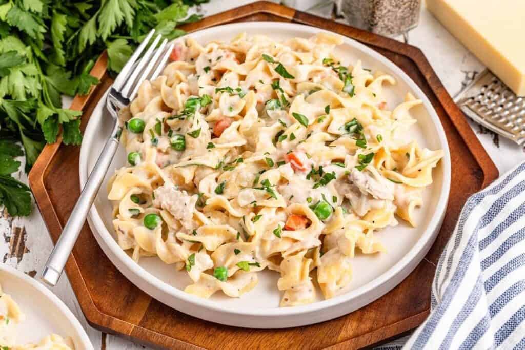 A plate of creamy chicken noodle casserole with peas and carrots, a classic from our favorite casserole recipes, is garnished with parsley and sits on a round wooden tray beside a fork.