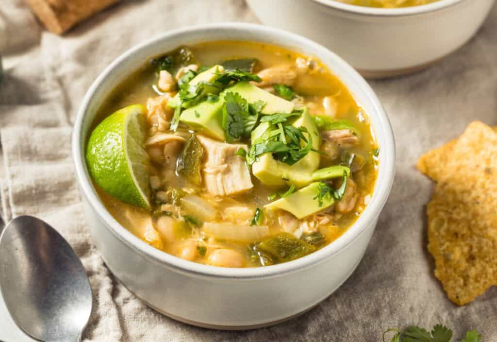 A bowl of white chicken chili topped with avocado, cilantro, and a lime wedge, with a spoon and tortilla chip nearby.