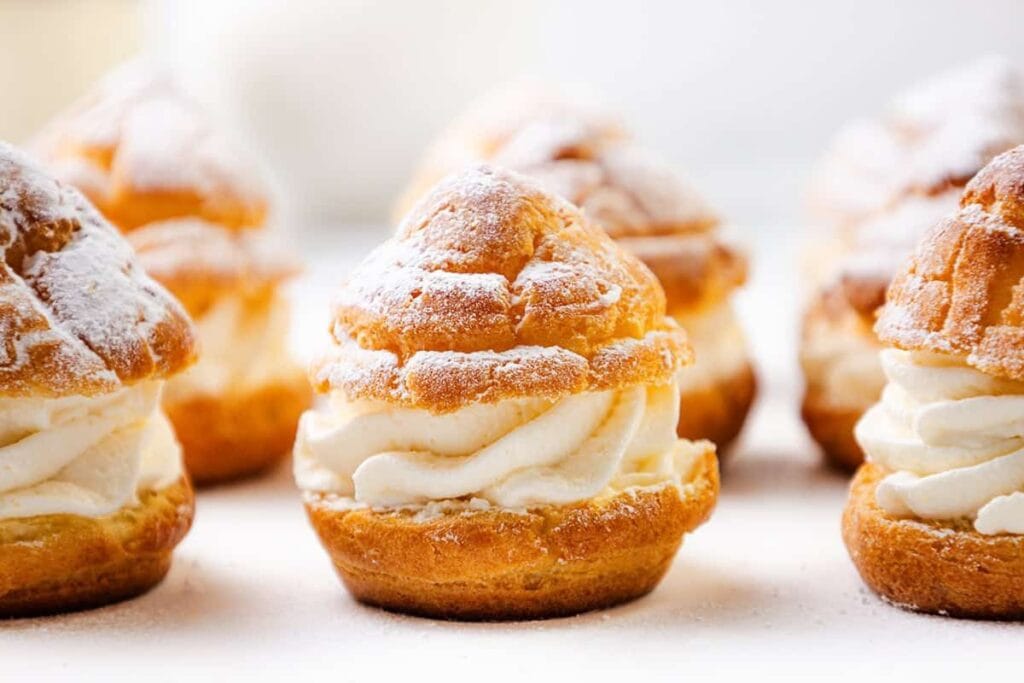 Cream puffs filled with whipped cream, dusted with powdered sugar, arranged on a light surface&mdash;perfect for adding a touch of elegance to your Bridgerton recipes collection.