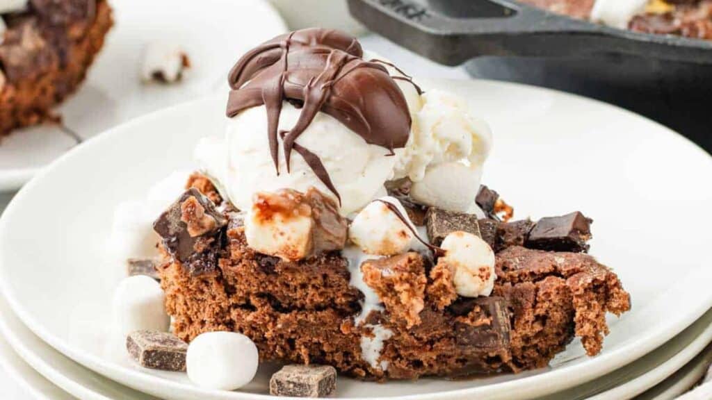 A slice of chocolate brownie topped with vanilla ice cream, mini marshmallows, chocolate chunks, and a drizzle of chocolate sauce on a white plate.