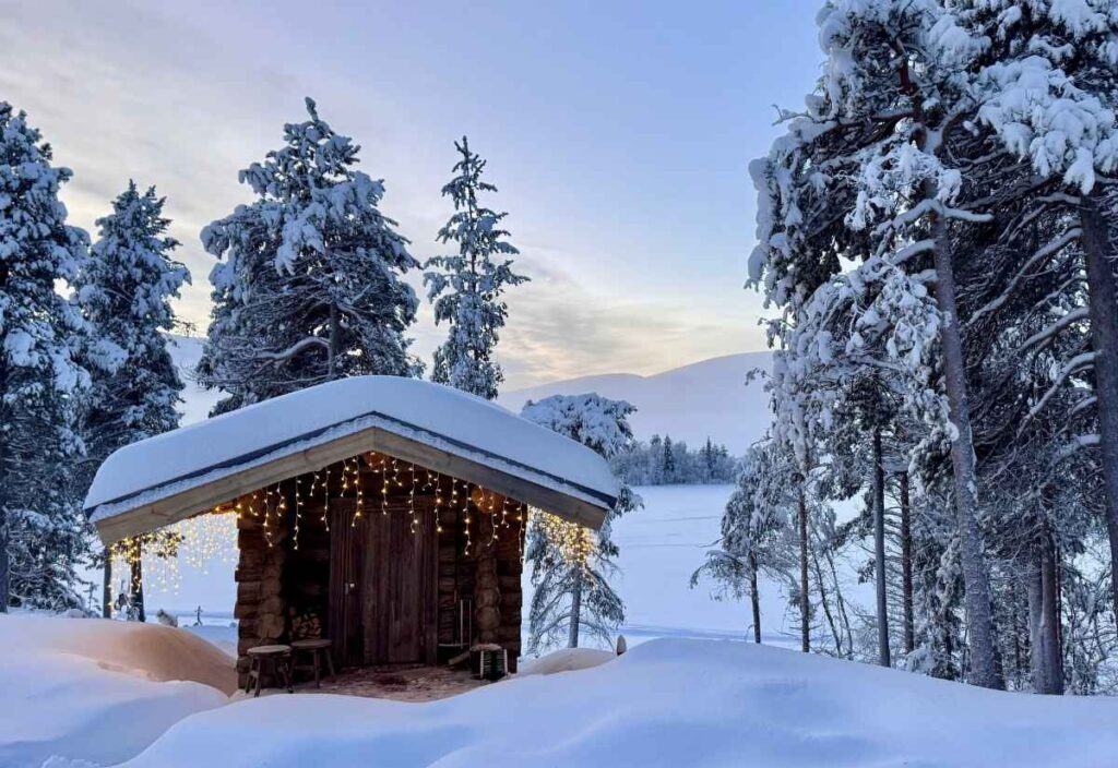 A small wooden cabin with string lights is surrounded by snow-covered trees and deep snowdrifts in a winter landscape at dusk.