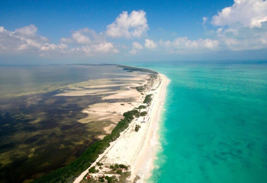 Aerial view of a long, narrow sandy beach bordered by green vegetation, with turquoise ocean on one side and shallow, darker tidal waters on the other under a partly cloudy sky.