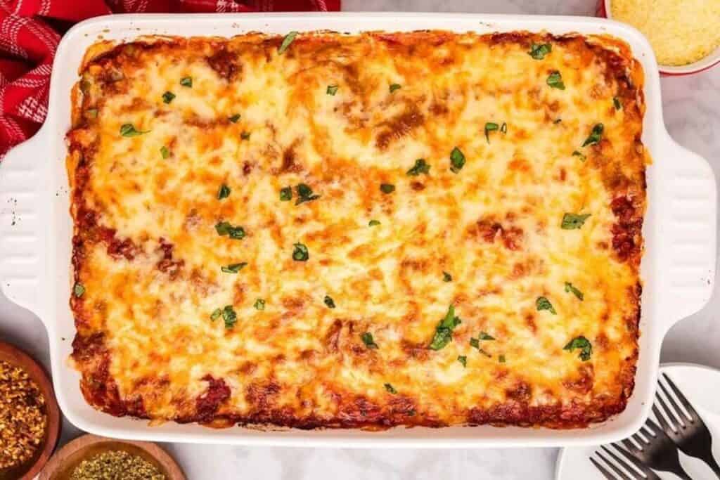 A baked lasagna in a white rectangular dish, topped with melted cheese and garnished with chopped herbs, sits on a countertop next to small bowls of spices&mdash;a classic favorite among casserole recipes.