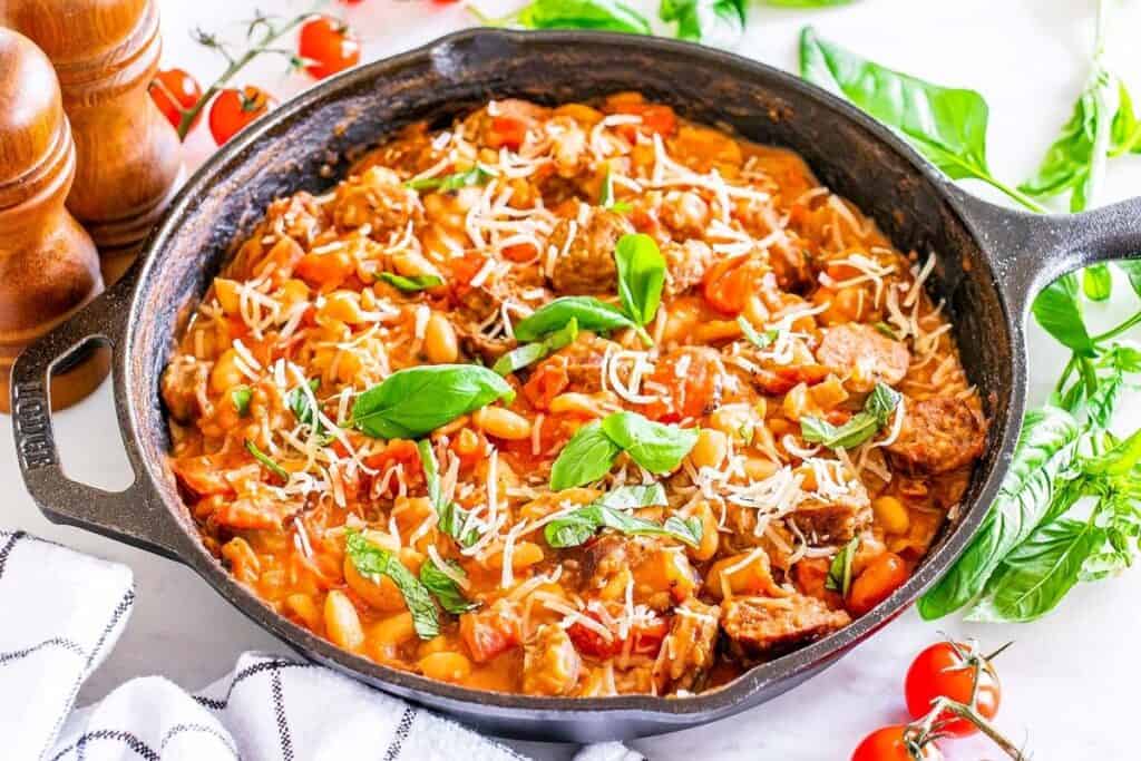 A cast-iron skillet filled with a sausage, rice, and tomato dish, garnished with basil and shredded cheese, sits on a white surface&mdash;one of those Simple 30 Minute Meals perfect for busy weeknights, with fresh herbs and tomatoes nearby.
