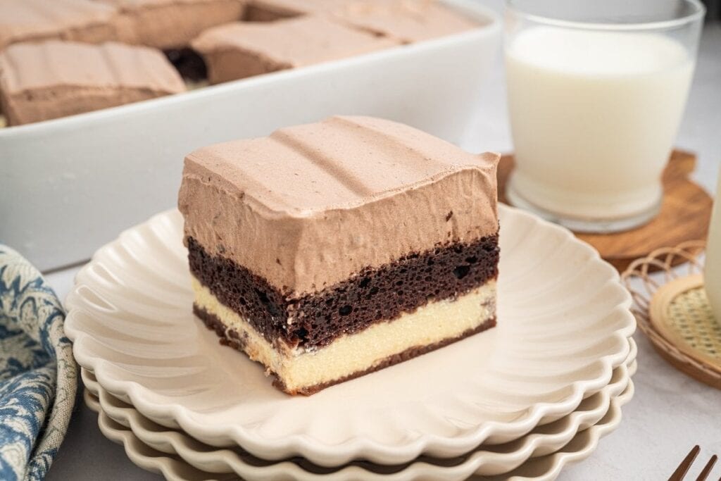A slice of layered chocolate and vanilla cake with chocolate frosting, a classic among easy desserts, sits on a stack of plates next to a glass of milk.