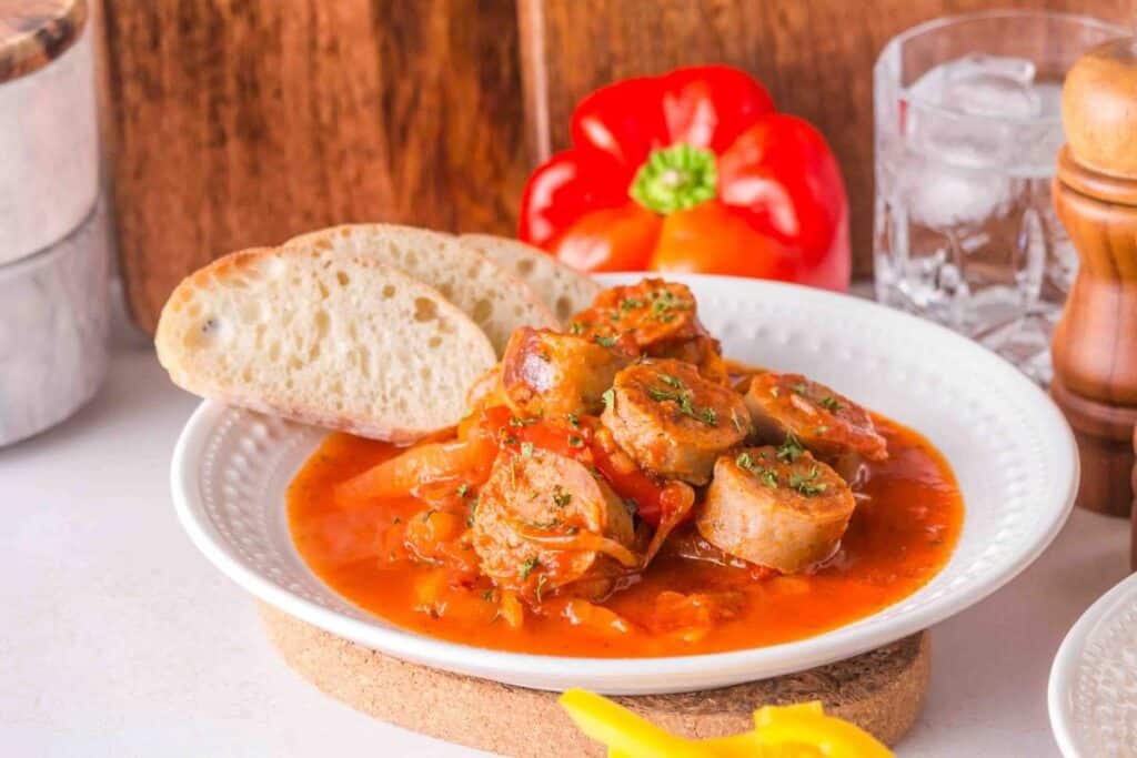 A plate of sliced sausage in tomato sauce with onions and herbs, served with two slices of bread—perfect for low effort dinners. A red bell pepper and a glass of ice water are in the background.