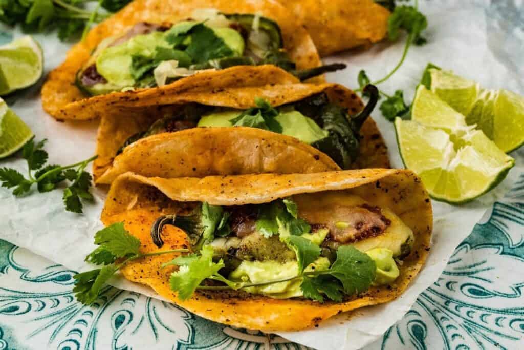 Three tacos filled with avocado, cheese, cilantro, and peppers are arranged on parchment paper with lime wedges on the side&mdash;perfect for Simple 30 Minute Meals.