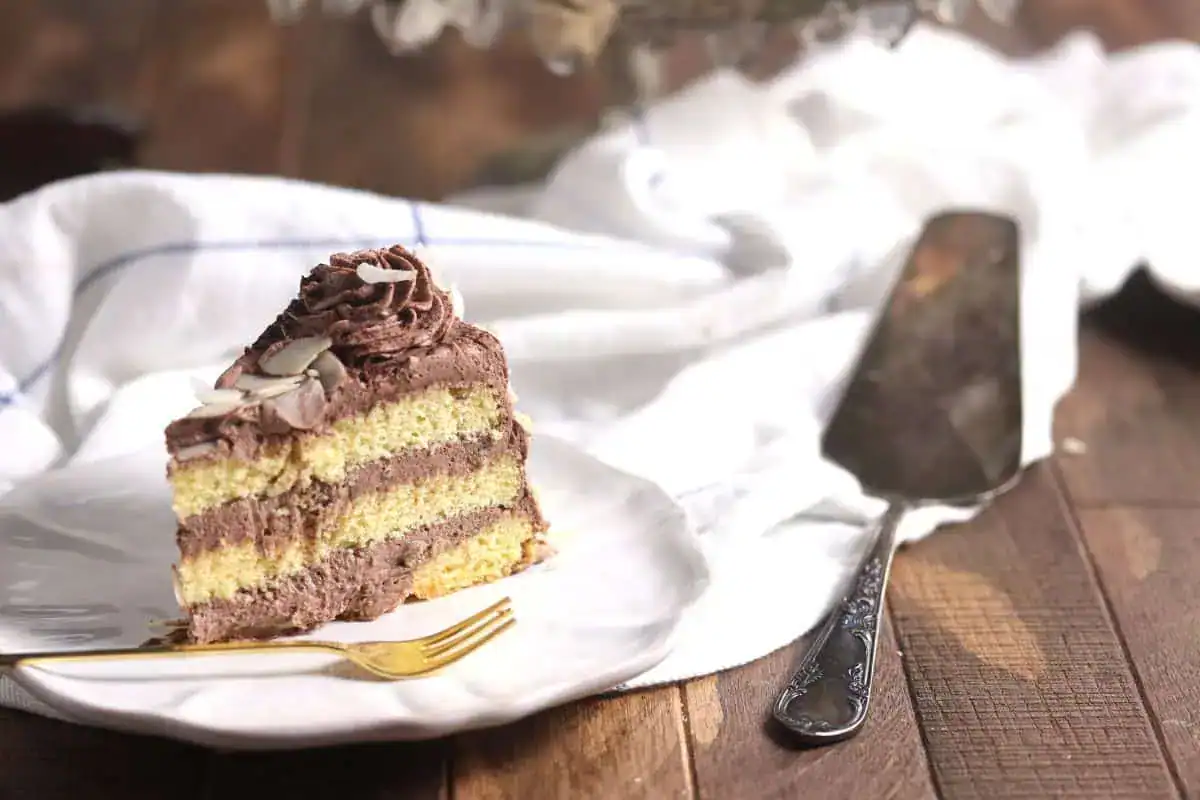A slice of layered yellow cake with chocolate frosting on a white plate, next to a gold fork and a cake server on a wooden table with a white cloth.
