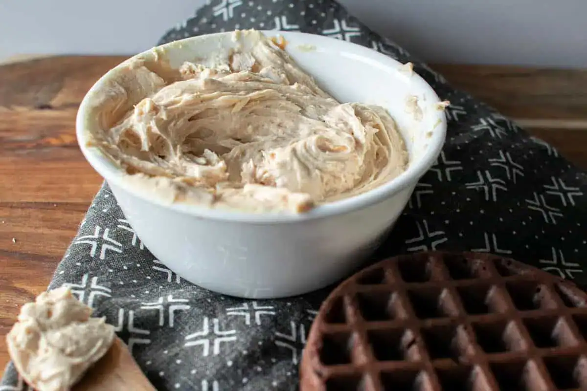 A white bowl filled with whipped peanut butter sits on a patterned cloth next to a chocolate waffle and a wooden spoon.