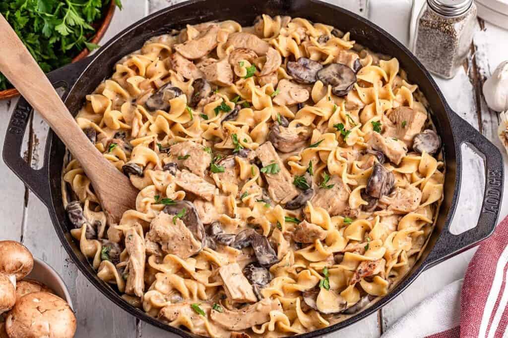A skillet filled with creamy chicken and mushroom pasta topped with fresh herbs, perfect for low effort dinners, with a wooden spoon resting inside.