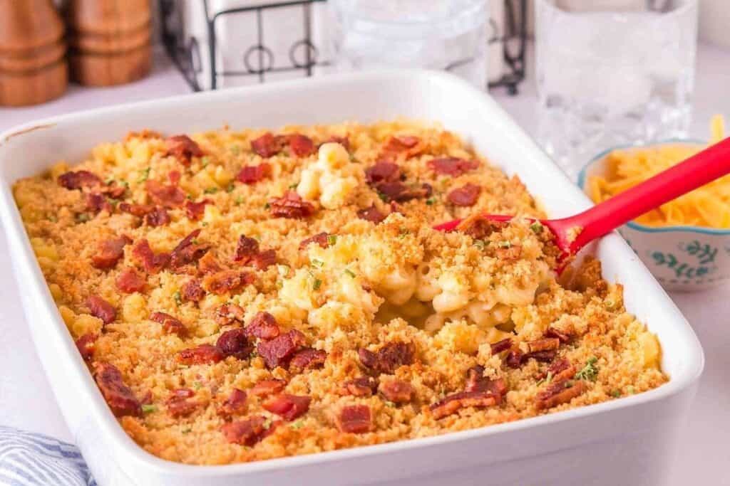 A Pasta Classics favorite: baked macaroni and cheese casserole topped with crispy breadcrumbs and bacon pieces, served in a white dish with a red serving spoon.