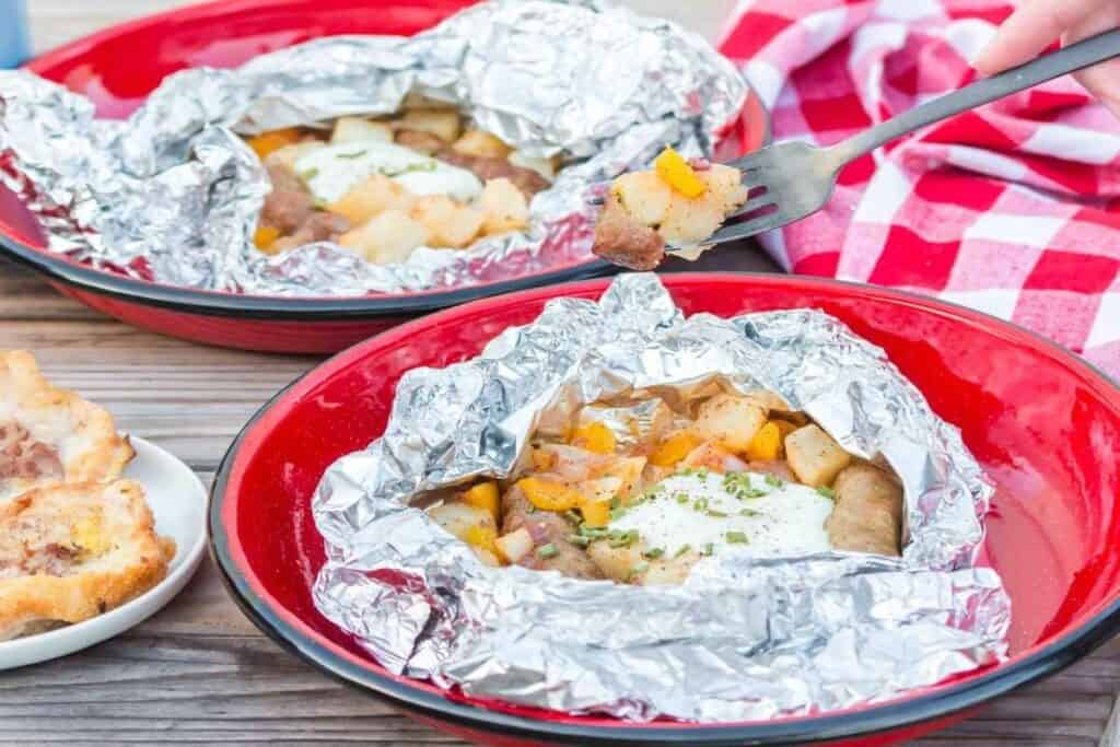 Two red plates with foil packets of sausage, potatoes, and vegetables topped with herbs make for tasty breakfast ideas. A hand holds a fork with a bite, and a red checkered napkin sits nearby.