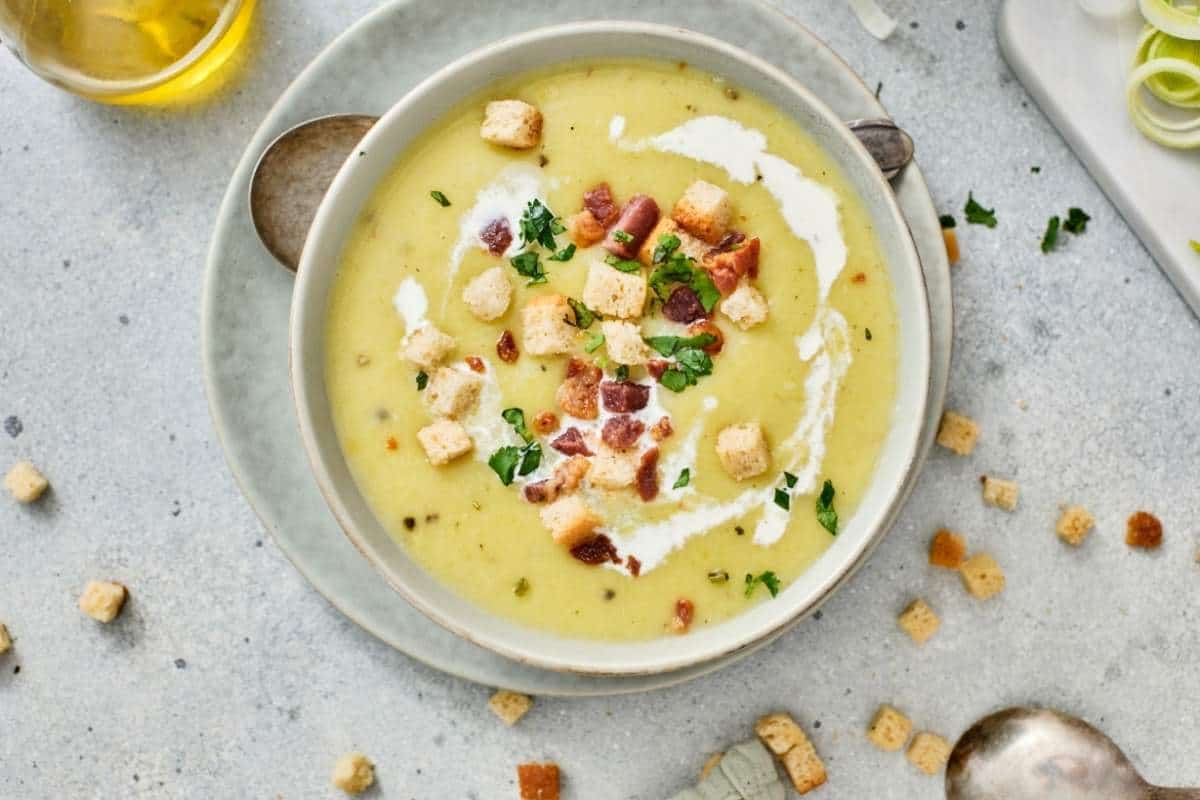 A bowl of Gordon Ramsay's Leek & Potato Soup is placed on a plate with a spoon nearby.