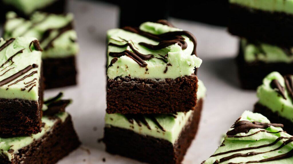 Stacked chocolate brownies topped with a layer of green mint frosting and drizzled with chocolate, surrounded by more brownies on a white surface.