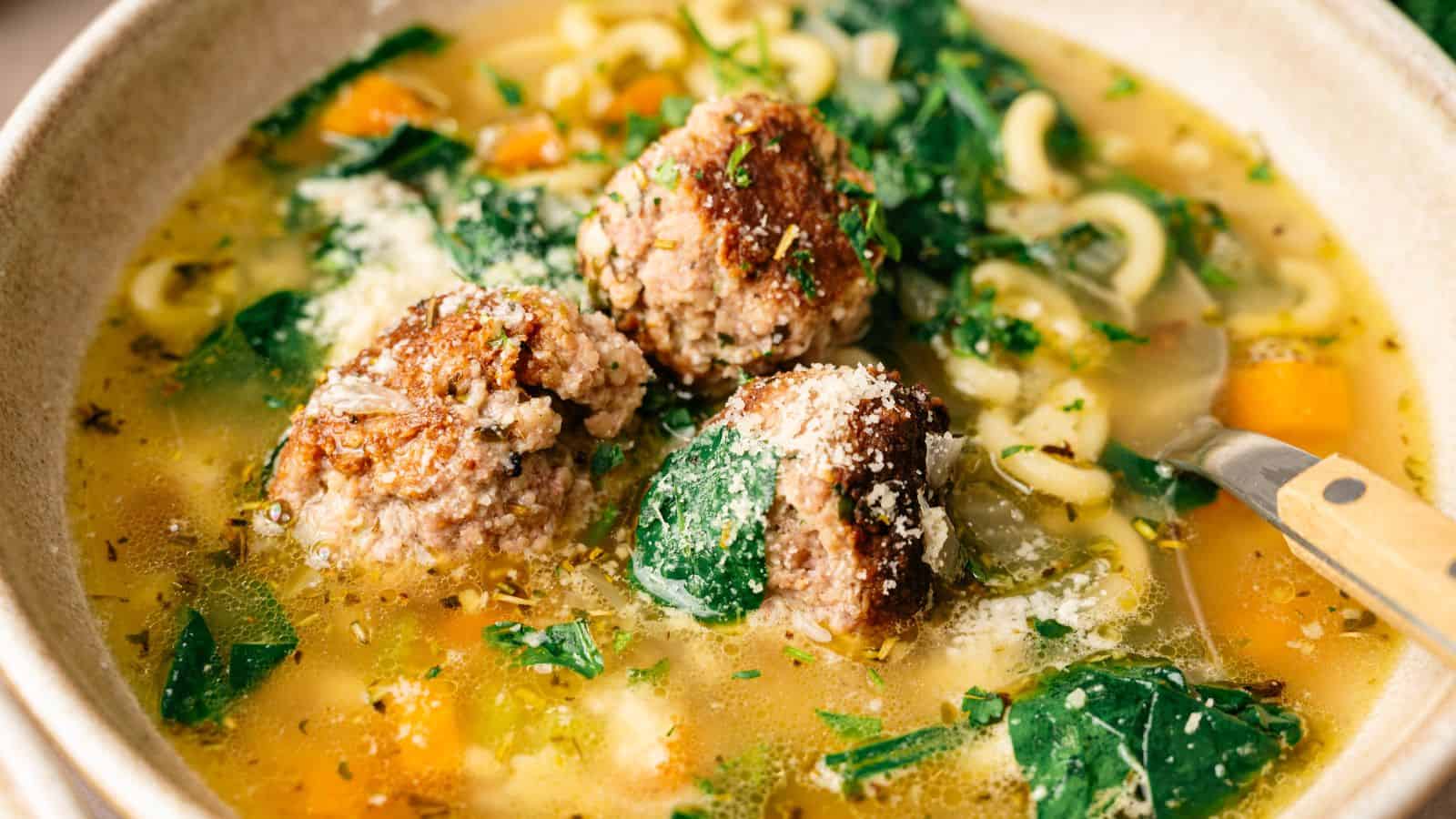 Bowl of soup with meatballs, spinach, pasta, and vegetables in broth, topped with grated cheese and herbs.