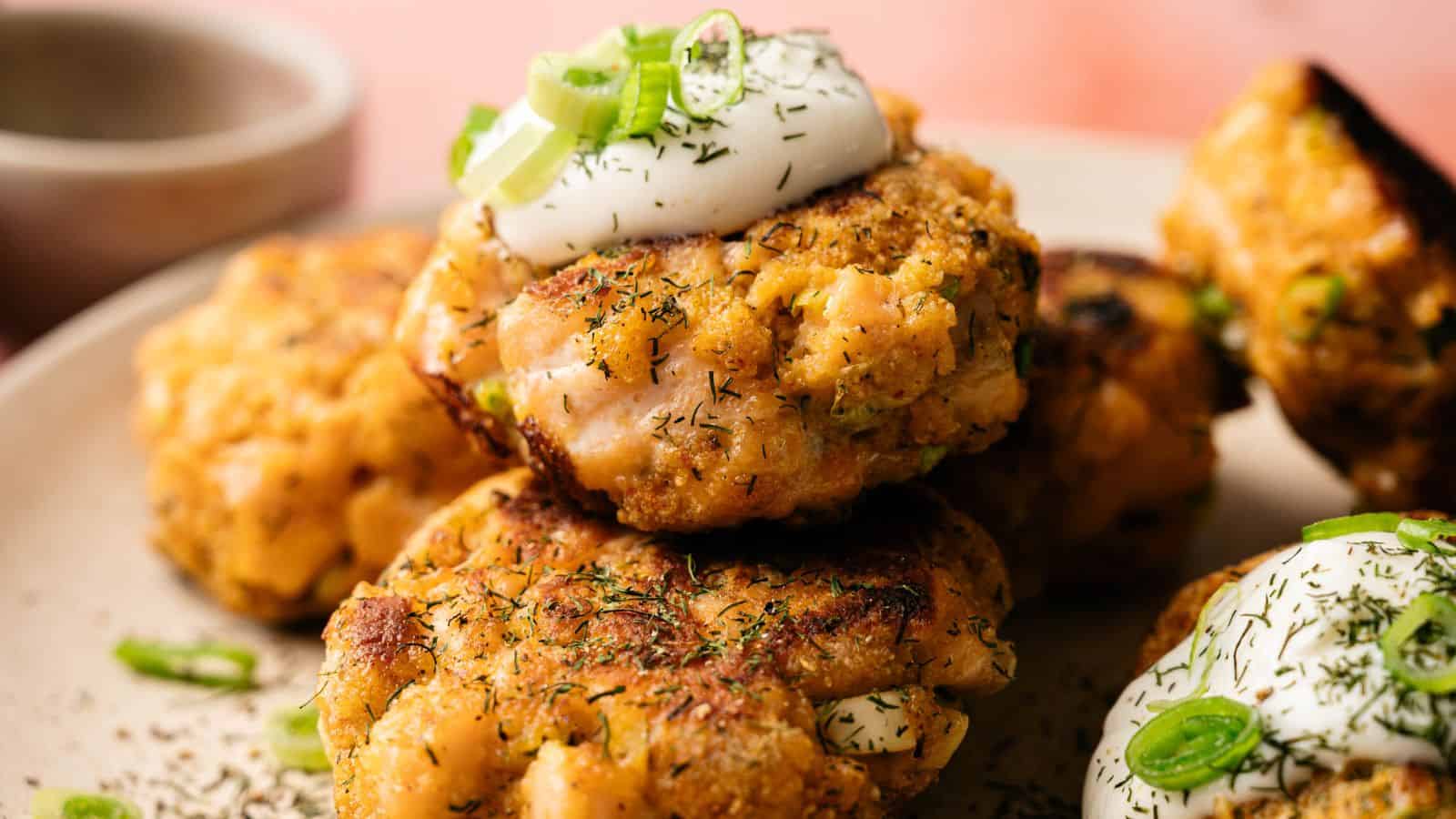 Close-up of golden-brown salmon patties topped with sour cream and chopped green onions, garnished with dried herbs on a plate.