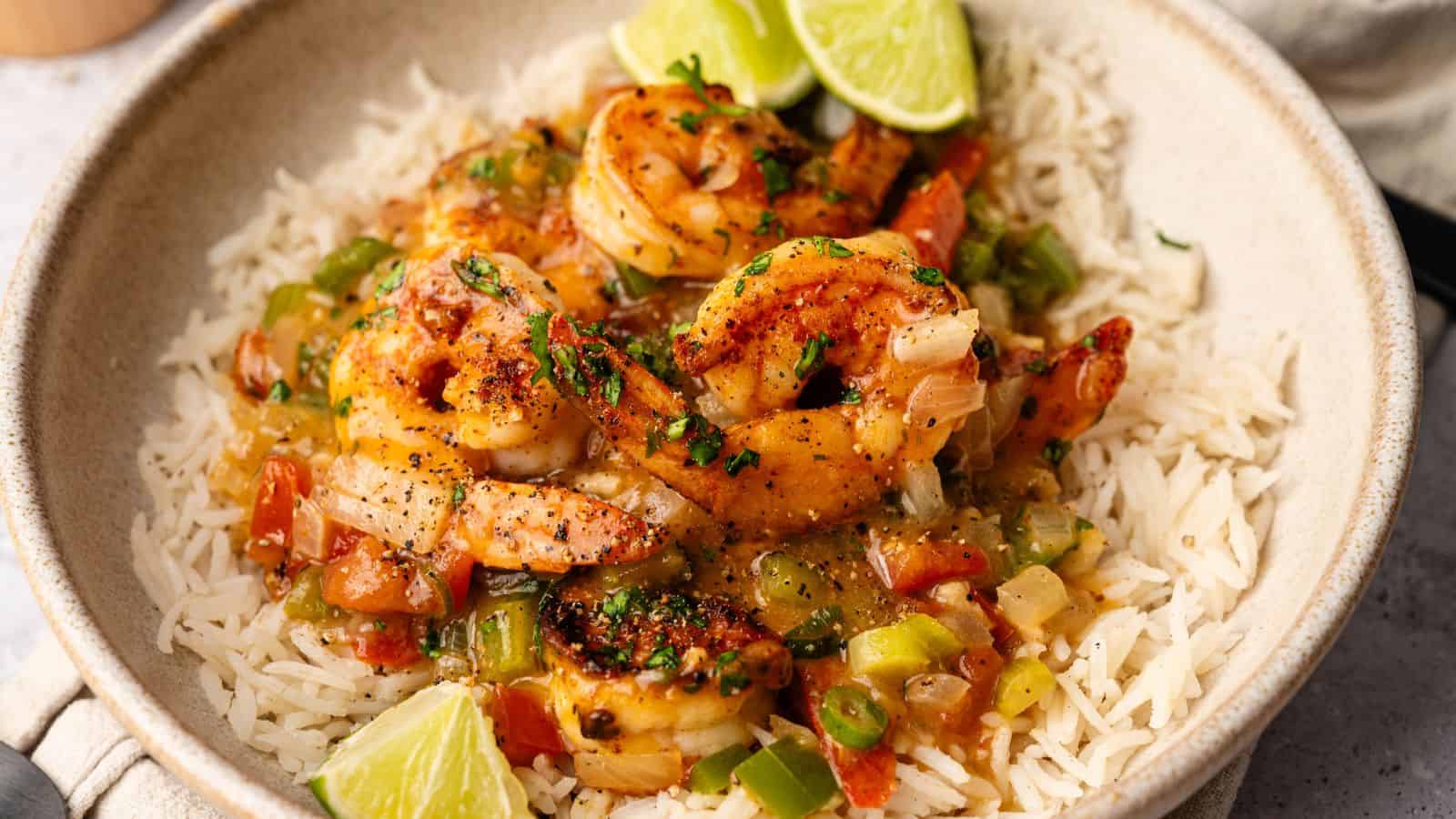 A bowl of white rice topped with cooked shrimp, diced tomatoes, green onions, and sauce, garnished with chopped herbs and lime wedges.