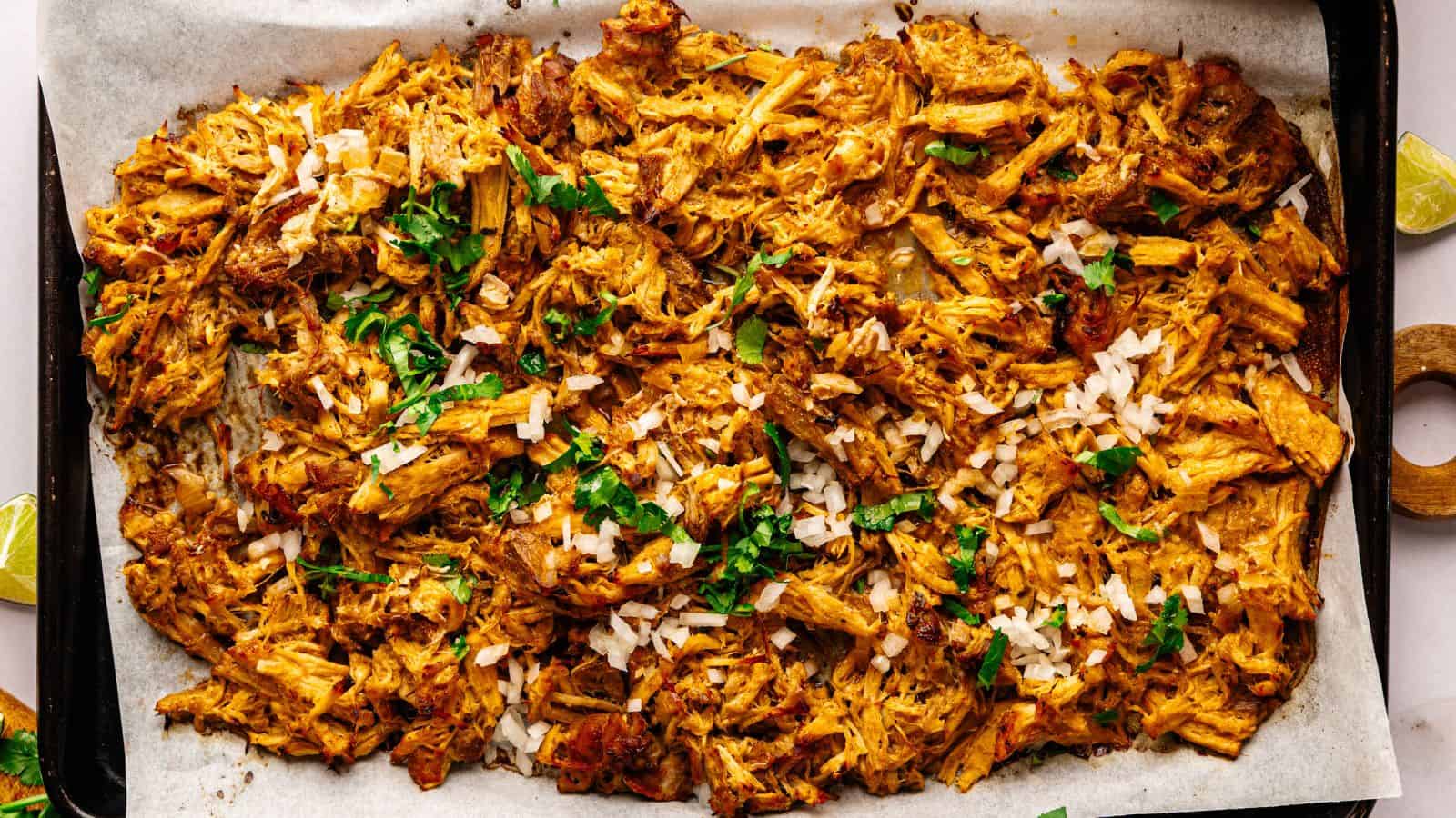 Shredded, seasoned meat spread on a parchment-lined baking sheet, topped with chopped onions and fresh cilantro.