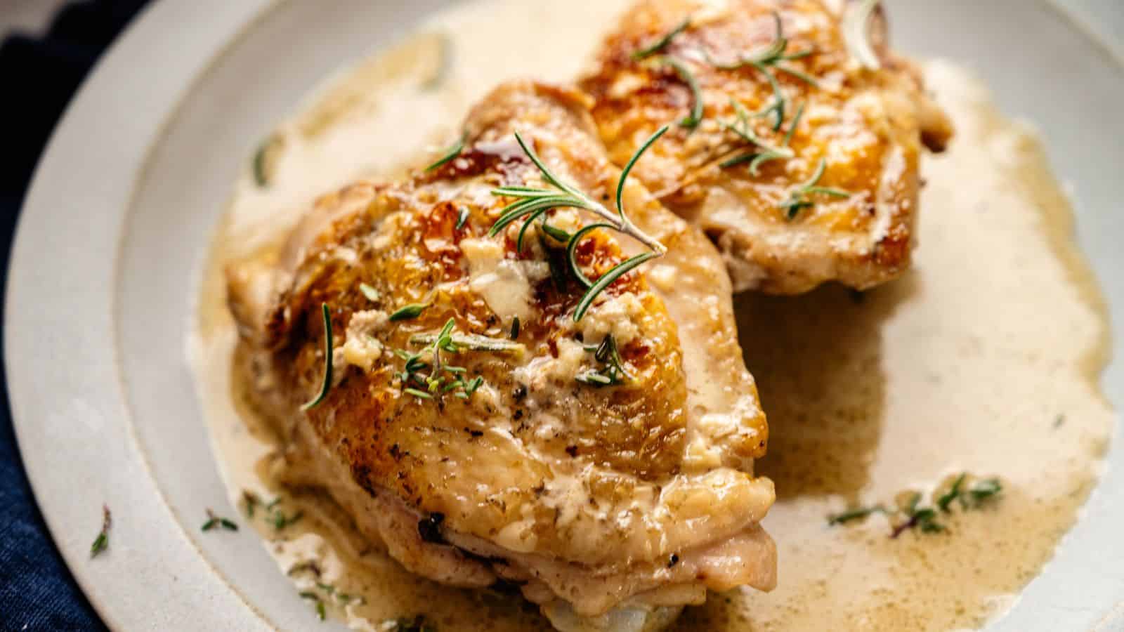 Two pieces of cooked chicken with crispy skin are served on a plate, topped with creamy sauce and garnished with sprigs of fresh herbs.