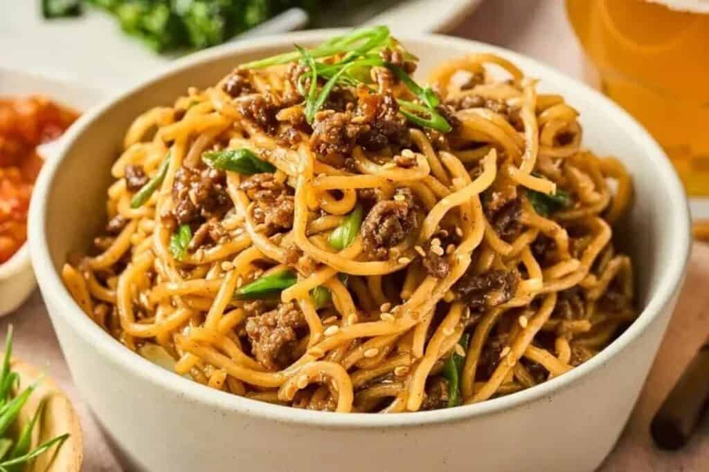 A perfect choice for weeknight dinners, this bowl of noodles is topped with minced meat, green onions, and sesame seeds, with tempting side dishes and a refreshing drink in the background.
