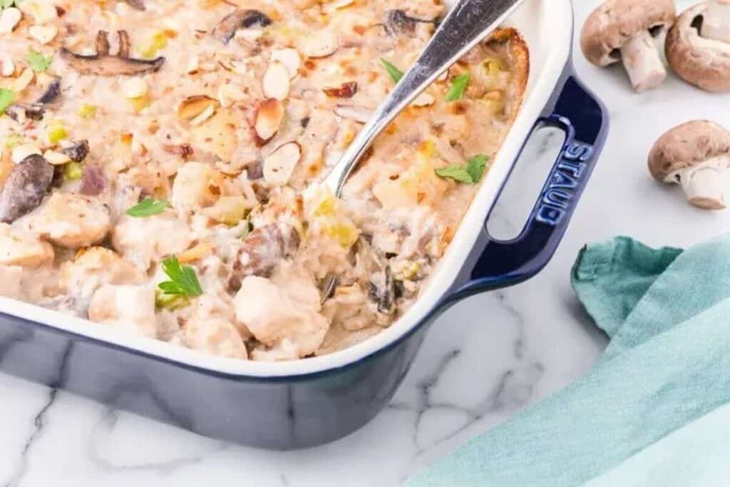 A baked casserole with chicken, mushrooms, and sliced almonds in a blue dish sits on a marble counter beside fresh mushrooms and a green cloth&mdash;perfect inspiration for your next casserole recipes.