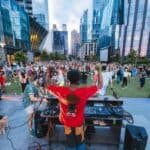 A DJ plays music on an outdoor stage as a large crowd gathers on a grassy plaza surrounded by modern glass buildings in the city.