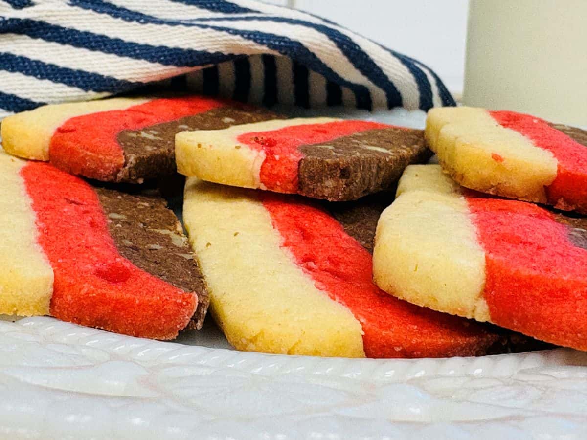 Rectangular cookies with red, white, and brown stripes are stacked on a white plate, with a striped cloth and a glass of milk in the background.