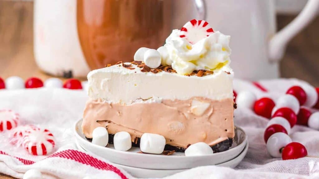 A slice of layered chocolate and cream dessert topped with whipped cream, peppermint candy, and mini marshmallows, surrounded by more candies and marshmallows on a plate.