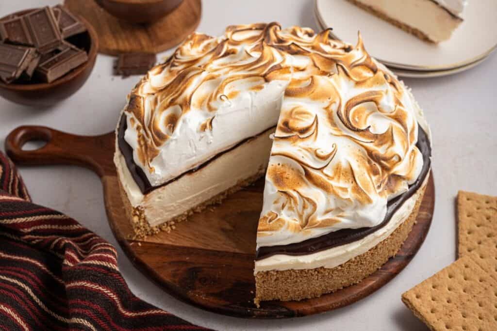 An easy dessert, this round s'mores cheesecake features a graham cracker crust, chocolate layer, and toasted meringue topping, beautifully displayed on a wooden board with a slice removed.