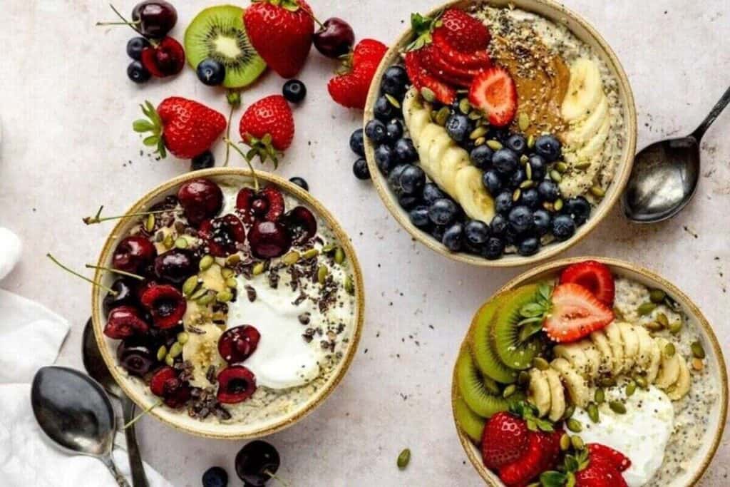 Three smoothie bowls topped with fresh fruits like strawberries, kiwi, banana, blueberries, and cherries, plus seeds and yogurt&mdash;perfect for those seeking tasty breakfast ideas. Arranged beautifully on a light surface with spoons.
