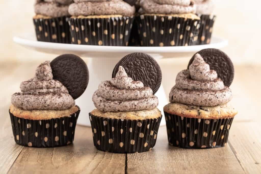 Three cupcakes with cookies and cream frosting and chocolate sandwich cookies on top are displayed in front of more cupcakes on a white cake stand&mdash;perfect for anyone looking for easy desserts.